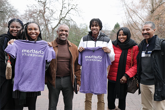 Two new Chatham University students smile, holding up purple ChathamU t-shirts and posing for a photo with their family members at New Student Registration Day